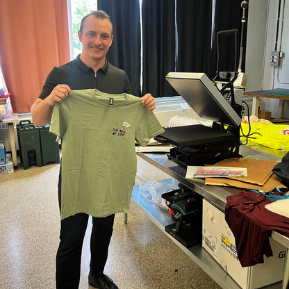 A man in a black polo shirt and black pants holds up a light sage green t-shirt with a small Box Of Life worm graphic on the left chest, standing in a workshop studio with a heat press machine and computer monitor on a workbench behind him.