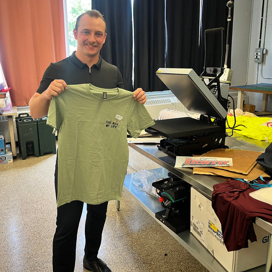 A man in a black polo shirt and black pants holds up a light sage green t-shirt with a small Box Of Life worm graphic on the left chest, standing in a workshop studio with a heat press machine and computer monitor on a workbench behind him.