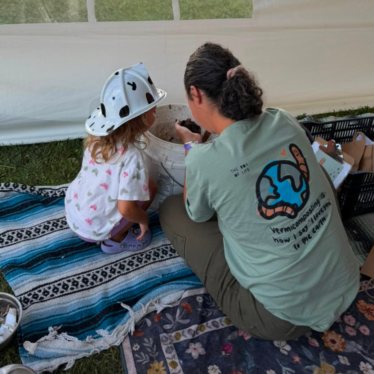 A woman and a young child sit on  blankets on the grass inside a white tent looking at a worm farm. The woman wears a light green T-shirt an earthworm wrapepd around the earth with the text 'vermicomposting is how say I love you to the earth'